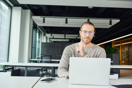 erfolgreicher Unternehmer mit stylischer Brille, der auf den Laptop schaut, während er an einem Geschäftsprojekt in der Nähe von Mobiltelefonen mit leerem Bildschirm am Arbeitsplatz in der modernen Umgebung des Coworking Office arbeitet