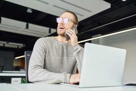 thoughtful businessman in eyeglasses looking away and talking on mobile phone while sitting near laptop on work desk, startup project planning in contemporary office space