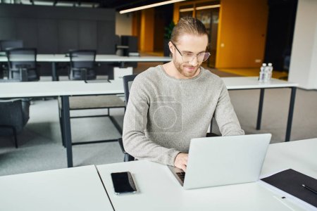 optimistic businessman in casual clothes and eyeglasses planning startup and typing on laptop near folder and smartphone with blank screen at workplace in contemporary coworking office