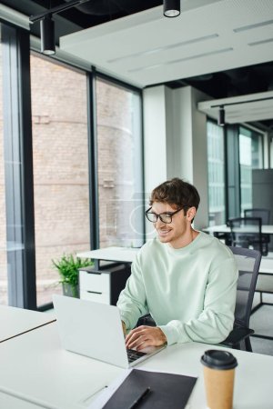 fröhlicher Unternehmer in stylischer Brille und lässiger Kleidung am modernen Arbeitsplatz sitzend und am Laptop in der Nähe von Ordnern und Coffee to go im modernen Büro vernetzt, erfolgreiches Geschäftskonzept