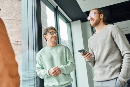 businessman in eyeglasses and stylish casual clothes standing with hand in pocket and smartphone near happy colleague and large window in modern office, concept of successful collaboration