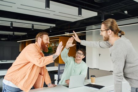 Photo for Overjoyed bearded and tattooed entrepreneur giving high five to business partners near laptop and takeaway drink in modern environment of coworking office, concept of successful collaboration - Royalty Free Image