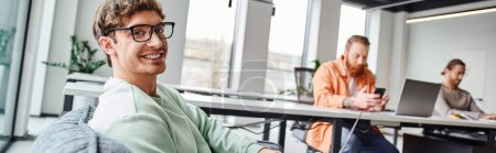 successful and optimistic team lead in stylish eyeglasses smiling at camera in modern office environment near colleagues working on blurred background, productive coworking concept, banner