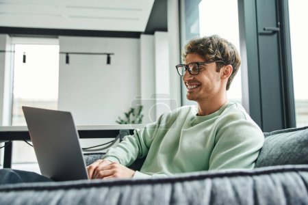 fröhlicher und erfolgreicher Geschäftsmann in stylischer Brille und lässiger Kleidung sitzt auf bequemer Couch in der Bürolounge des Coworking Space und vernetzt sich am Laptop, Geschäftsidee-Konzept