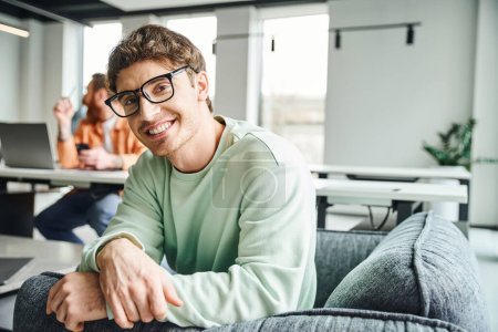 glücklicher Unternehmer in stylischer Brille und lässiger Kleidung im Sessel sitzend und lächelnd in die Kamera neben Kollegen, die auf verschwommenem Hintergrund im modernen Büro arbeiten, produktives Coworking-Konzept