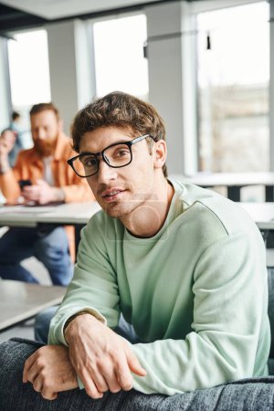 positive and ambitious entrepreneur in eyeglasses and casual clothes looking at camera while his business colleague working on blurred background in contemporary office, productive coworking concept