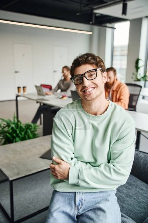 cheerful businessman in eyeglasses and casual clothes sitting with folded arms and looking at camera while colleagues working near laptops on blurred background, productive coworking concept