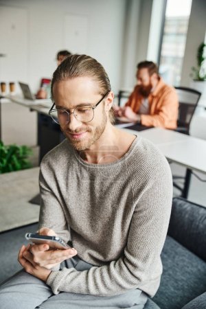 Lächelnder Geschäftsmann mit Brille und lässiger Kleidung, Vernetzung auf dem Smartphone in der Nähe von Kollegen, die an verschwommenem Hintergrund arbeiten, erfolgreiches Unternehmertum-Konzept