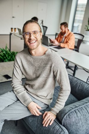 glücklicher Unternehmer mit stylischer Brille, der in einem modernen Coworking-Umfeld in der Nähe von Kollegen mit verschwommenem Hintergrund in die Kamera blickt, erfolgreiches Unternehmertum-Konzept