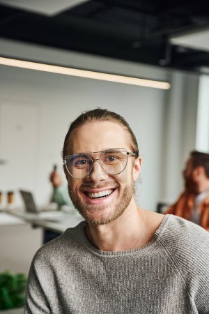 professional headshot of handsome and overjoyed businessman in stylish eyeglasses smiling at camera on blurred background in modern coworking office, successful entrepreneurship concept