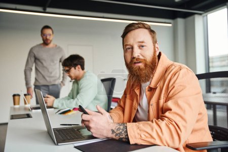 bearded and tattooed businessman in stylish casual clothes holding smartphone and looking at camera near laptop and colleagues working on blurred background in contemporary coworking space