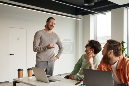 positiver Unternehmer mit stylischer Brille, der mit der Hand auf der Hüfte steht und mit dem Finger zeigt, während er mit seinem Team im modernen Coworking-Büro an Laptops sitzend Geschäftsprojekte diskutiert