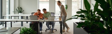 entrepreneur in casual clothes standing next to colleagues working on new business project near computers in modern office environment with high tech interior and natural plants, banner