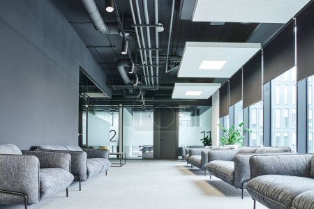 spacious waiting area with grey and comfortable sofas, large windows and green plants in contemporary office environment, workspace organization concept