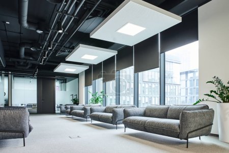 amplio salón de espera con sofás grises y cómodos, plantas naturales verdes y grandes ventanales en la moderna oficina de coworking, concepto de organización del espacio de trabajo