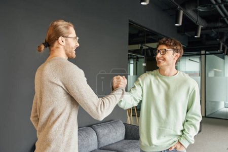 Photo for Cheerful and stylish entrepreneurs in eyeglasses and casual clothes shaking hands and confirming agreement in lounge of modern coworking office, successful collaboration concept - Royalty Free Image