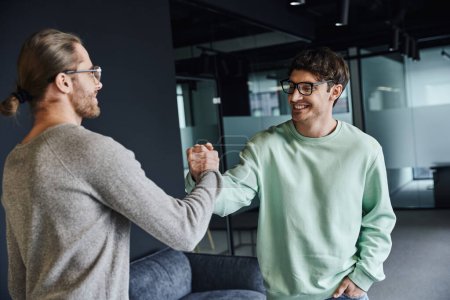 stylish and joyful business partners in eyeglasses and casual clothes confirming agreement and shaking hands in modern lobby of coworking office space, successful collaboration concept