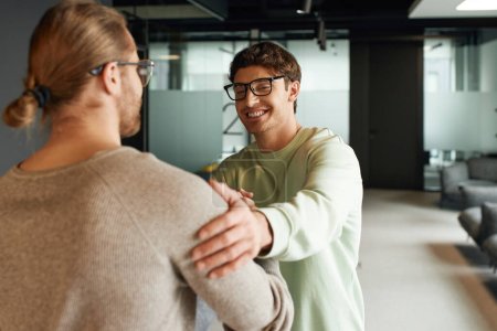 happy businessman in casual clothes and eyeglasses shaking hands and touching shoulder of colleague while confirming deal in lounge of modern coworking space, successful partnership concept