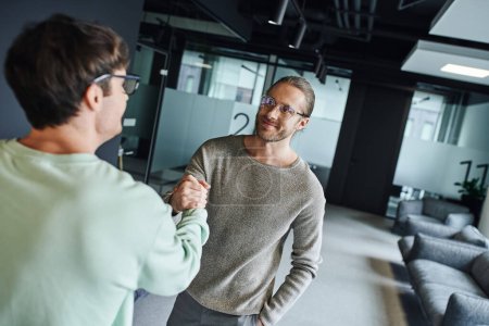 pleased businessman in eyeglasses and stylish casual clothes shaking hands with partner while closing deal in lounge of contemporary coworking office, successful collaboration concept