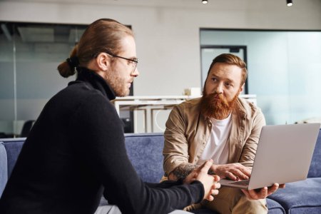 bearded businessman showing startup project on laptop to serious entrepreneur in black turtleneck and eyeglasses while sitting on couch in contemporary office, business partnership concept