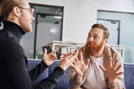 entrepreneurs gesturing and discussing startup project in modern office, bearded tattooed businessman with colleague in black turtleneck, business collaboration concept