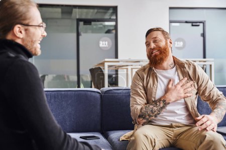 grateful, bearded and tattooed entrepreneur touching chest during conversation with business partner on couch in modern office environment, business collaboration concept