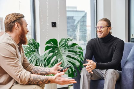 bearded and tattooed entrepreneur holding smartphone and discussing business project with smiling colleague near green plant in contemporary office, business collaboration concept