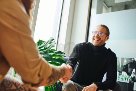 excited businessman in eyeglasses and black turtleneck shaking hands with tattooed businessman on blurred foreground in modern office, successful partnership concept