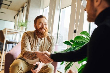 cheerful, bearded and tattooed businessman shaking hands with ambitious entrepreneur in black turtleneck while sitting in contemporary office, successful partnership concept