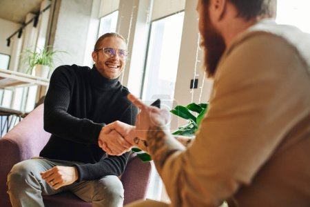 Photo for Happy entrepreneur in eyeglasses and black turtleneck shaking hands and closing deal with tattooed businessman pointing with finger on blurred foreground, successful partnership concept - Royalty Free Image