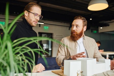 bearded and tattooed architect showing building model to thoughtful colleague in eyeglasses and black turtleneck near blurred plant in contemporary design studio, architecture and business concept