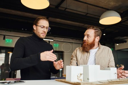 architect in eyeglasses and black turtleneck gesturing near building model while discussing startup project with bearded colleague in design studio, architecture and business concept