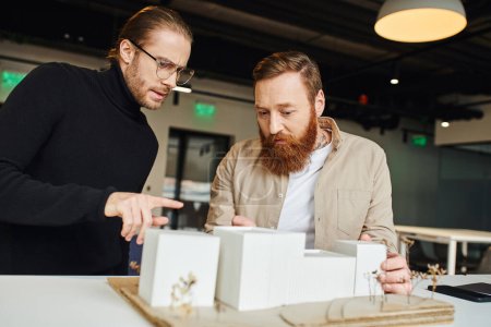 serious architect in eyeglasses and black turtleneck pointing at building model and talking to bearded colleague working in contemporary design studio, architecture and business concept