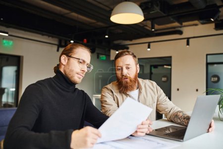 entrepreneur in black turtleneck and eyeglasses planning business project and working with papers near bearded and tattooed colleague sitting near computer in contemporary office environment