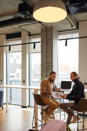 businessman in eyeglasses and black turtleneck looking at charts with bearded and tattooed colleague while sitting at laptop with blank screen and planning startup in modern office environment