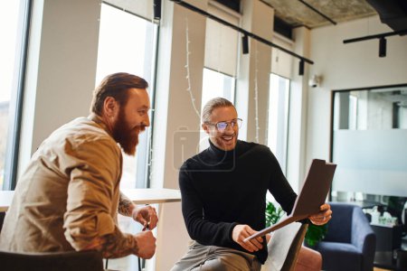 cheerful entrepreneur in black turtleneck and eyeglasses looking at folder with business project near bearded colleague in modern office, productivity and collaboration concept