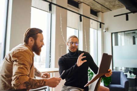 Inspirierter Geschäftsmann mit Brille und schwarzem Rollkragen zeigt Ordner mit Startup-Planung dem bärtigen Kollegen in zeitgenössischem Coworking-Büro, Produktivität und Kollaborationskonzept