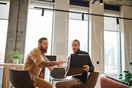 bearded and tattooed businessman pointing with pen at folder with startup project near colleague while sitting in contemporary office with large windows, productivity and collaboration concept