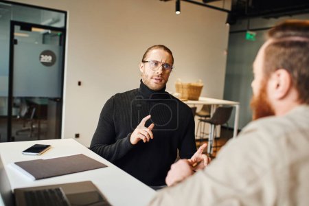 confident entrepreneur in black turtleneck and eyeglasses gesturing and talking to colleague near laptop and mobile phone, serious businessmen discussing startup project in modern office space
