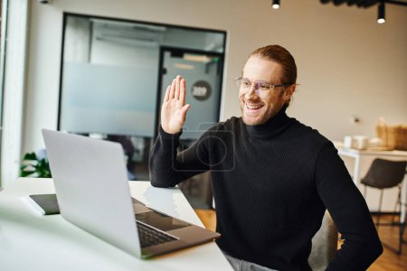 Lächelnder Unternehmer in schwarzem Rollkragen und Brille winkt bei Videokonferenz am Laptop am Arbeitsplatz in modernem Coworking-Büro, Business-Lifestyle-Konzept
