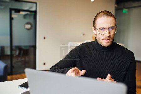 serious entrepreneur in black turtleneck and eyeglasses having video call on laptop, talking and gesturing in coworking environment of modern office, business lifestyle concept
