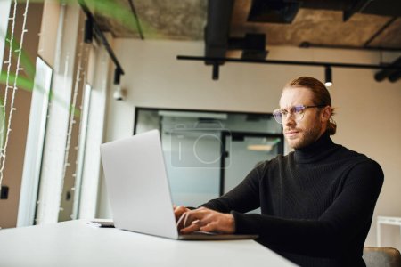 Konzentrierter Geschäftsmann mit schwarzem Rollkragen und Brille, der in Laptopnähe denkt und an einem Startup-Projekt in moderner Büroumgebung arbeitet, Business-Lifestyle-Konzept