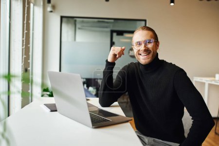 happy entrepreneur in black turtleneck and eyeglasses sitting near laptop on work desk and looking at camera in coworking environment in modern office, business lifestyle concept