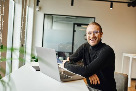homme d'affaires joyeux et élégant en col roulé noir et lunettes regardant la caméra tout en étant assis près d'un ordinateur portable avec les bras croisés dans un environnement de bureau moderne, concept de style de vie d'affaires