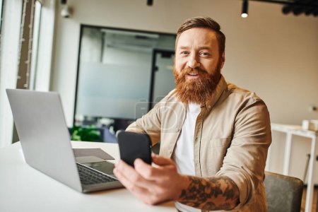 happy tattooed and bearded entrepreneur holding smartphone and smiling at camera while sitting near laptop in contemporary coworking office, business lifestyle concept