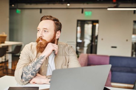 professional headshot of serious, bearded and tattooed businessman planning startup project while sitting near laptop and looking away in modern office environment, business lifestyle concept