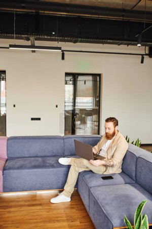 longitud completa de hombre de negocios tatuado y barbudo en ropa casual elegante trabajando en el ordenador portátil mientras está sentado en un cómodo sofá cerca de teléfono inteligente en el salón de la oficina moderna, concepto de estilo de vida de negocios
