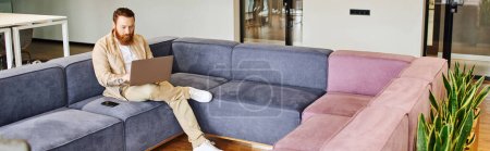 full length of stylish, bearded and tattooed entrepreneur sitting on comfortable couch near smartphone and working on computer in lounge of contemporary office, business lifestyle concept, banner