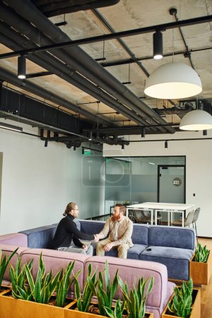 business partners in stylish casual clothes shaking hands and confirming agreement while sitting on cozy couch in modern office lounge with high tech interior, partnership and success concept