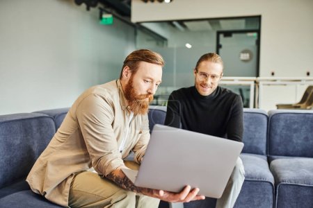 bearded and tattooed entrepreneur showing laptop with business project to pleased colleague in lounge of contemporary office environment, business partnership concept